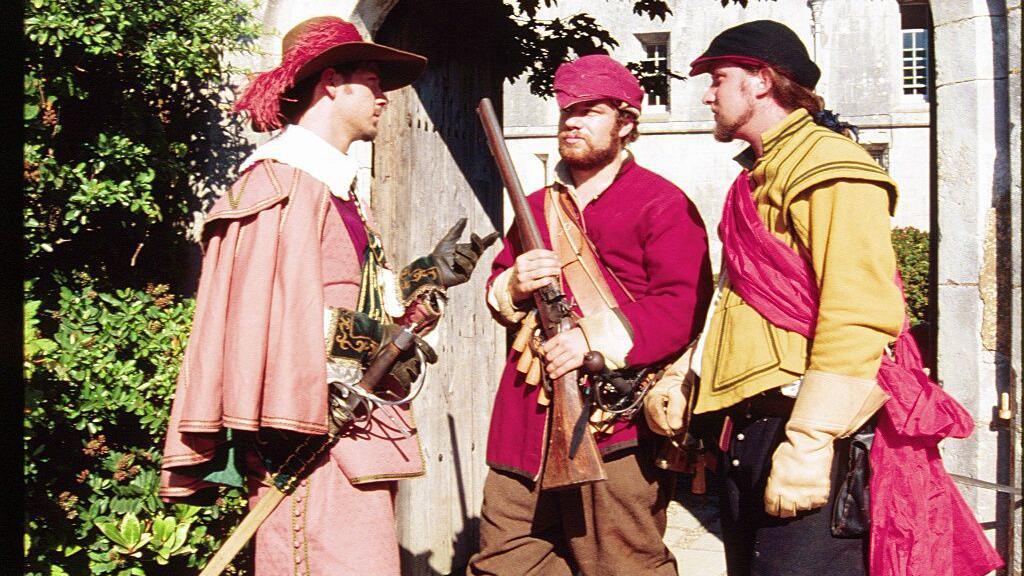 Three men wearing medieval attire and holding fake guns and swords, standing outside an old building.