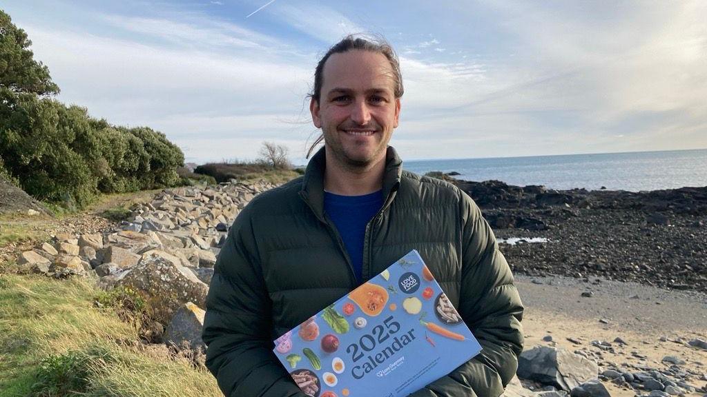 Douglas Button from Guernsey Waste holding the 2025 Love Food, Hate Waste Calendar, in front of a beach