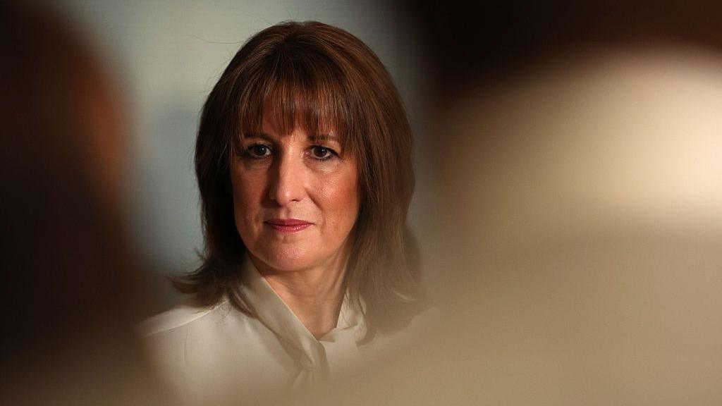 Rachel Reeves, dressed in a white blouse, can be seen in focus between the heads of an audience