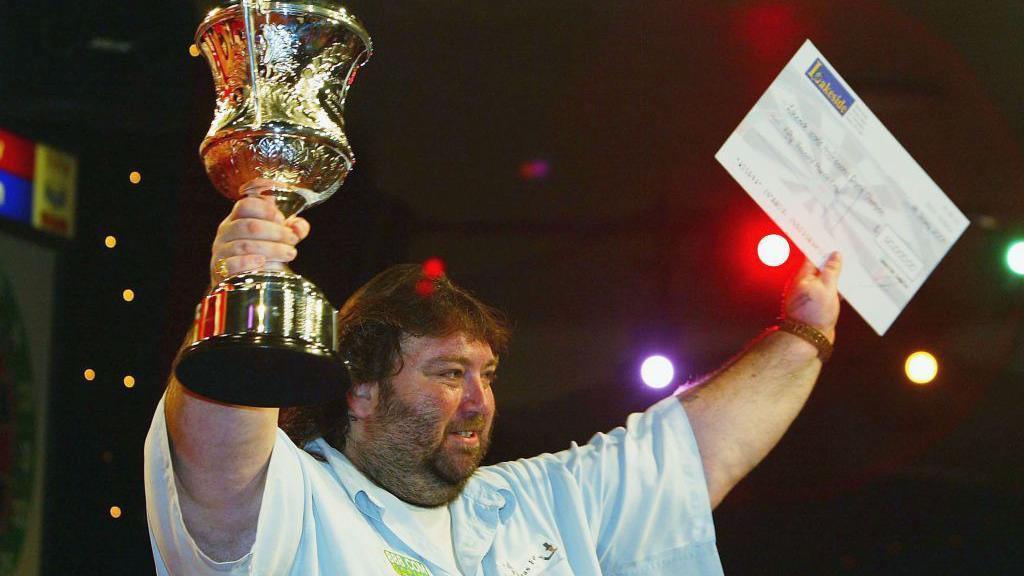 Andy Fordham holding a trophy and a large cheque. He is wearing a white top