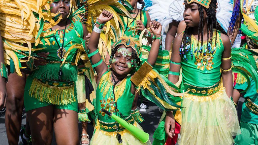 girls dressed in green tutu skirts join the parade, smaller girl in the middle smiles and raises her arms in the air