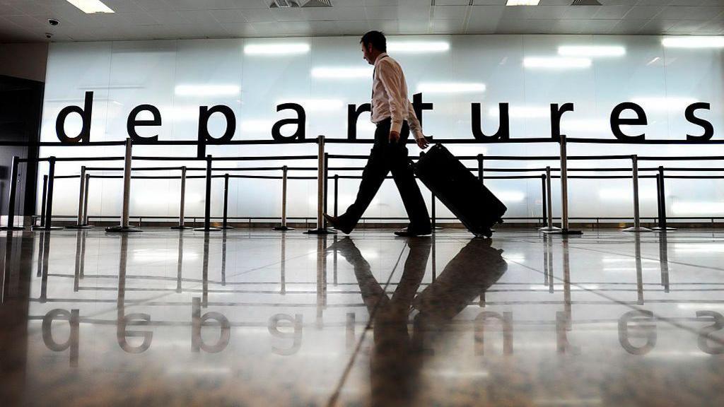 A man walking along through departures at Glasgow airport, carrying a wheeled trolley bag.