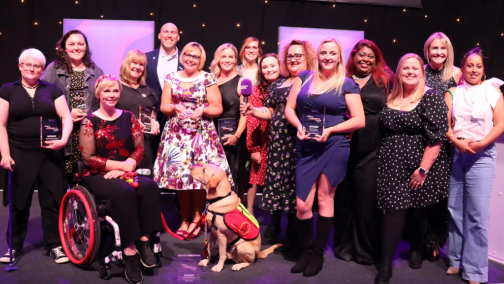 The winners of the BBC Bristol Make a Difference Awards 2024 line up for the camera in a large group. There is a dark wall with bright lights on it in the background