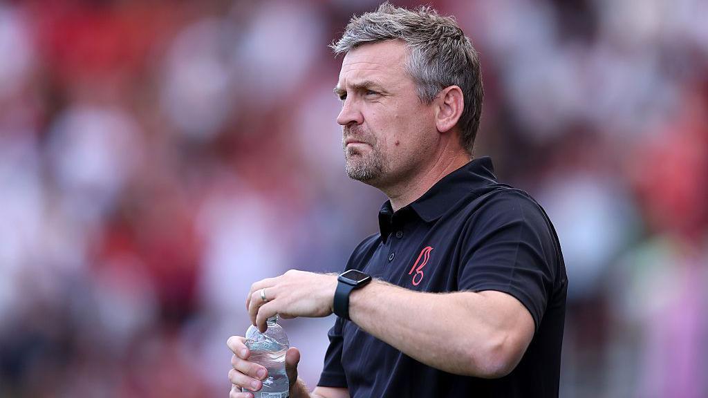 Bristol City boss Michael Skubala in a black short-sleeved polo shirt and holding a bottle of water on the touchline