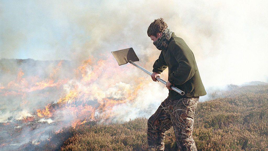 A man uses a spade to manage a fire on a moorland. He is wearing a green fleece and cargo trousers.