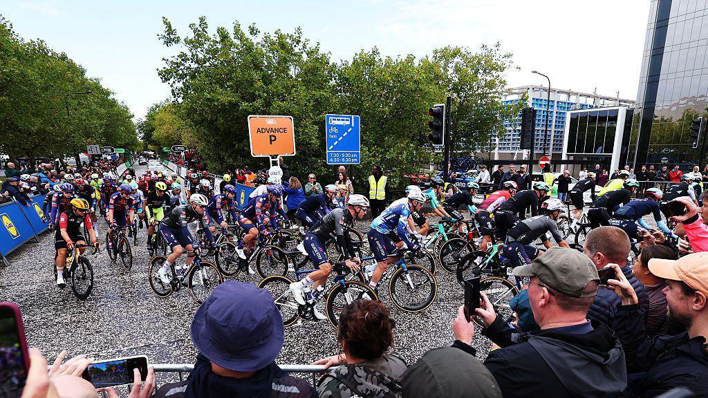 A swarm of cyclists speed round a bend as onlookers try to take video and photos on their phones. There are mirrored glass buildings behind them.