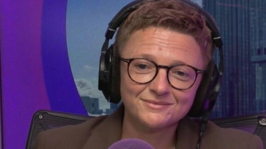 Bev Craig, with short cropped hair and glasses, smiles as she sits behind a microphone wearing headphones