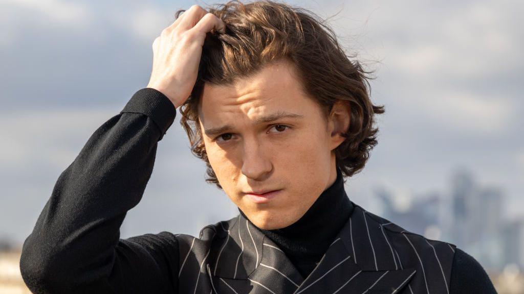 Tom Holland wearing a sharp pinstripe black suit runs his fingers through his hair and stares intently at the camera.