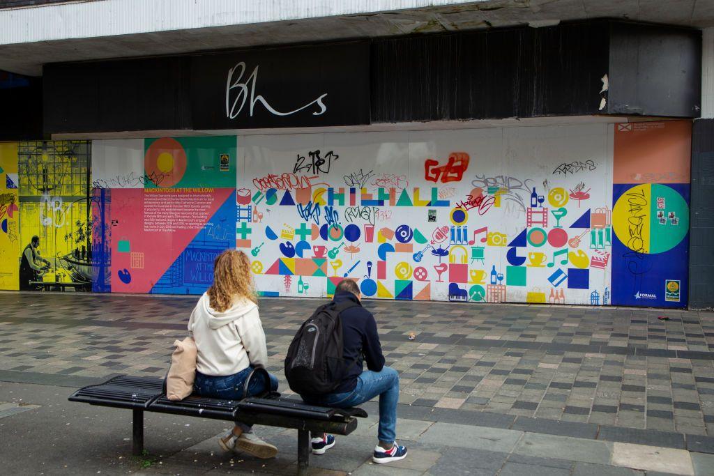 A man and woman sit on a bench in front of a former BHS store. The windows of the store have been covered with artwork, but it has been defaced by graffiti. There is litter on the pavement. 