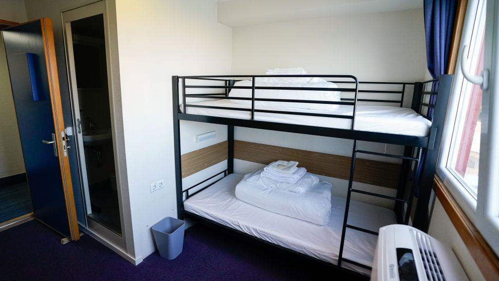 A basic room with a bunk bed and clean sheets in a pile on top of the beds. There is a radiator on the wall and a bin by the bed frame. There is a window on the wall