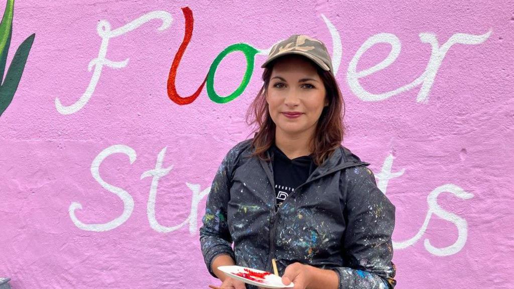 Madeleine Pires holds a paintbrush and palette of red paint, wearing a paint splattered dark waterproof jacket, black T-shirt and a camouflage design baseball cap. She stands in front of the words 'flower streets' which are set against a pink background. The letters 'l' and 'o' have been painted in red and green respectively.