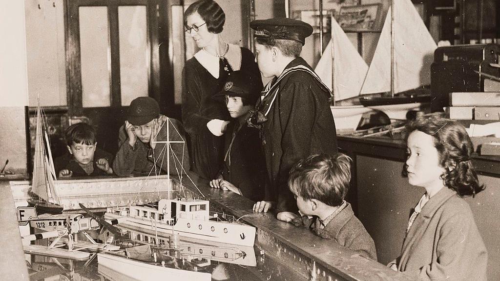 Gelatin silver print photograph. Young boys, some wearing caps and one wearing a sailor suit, look at a toy boat display as an adult woman wearing a big collared dress and glasses keeps watch. 