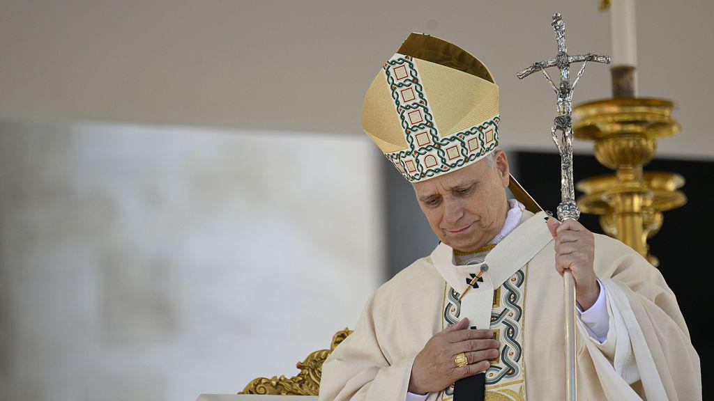Pope Leo wears cream papal garments embroidered with gold, a gold hat and ring, and holds a silver crucifix.