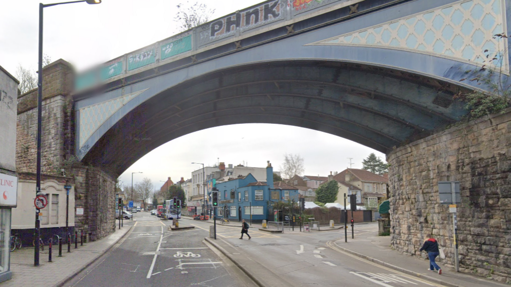 A view of a railbridge - painted blue - over a main route. There are buildings and people crossing the road. There are traffic lights set to green.