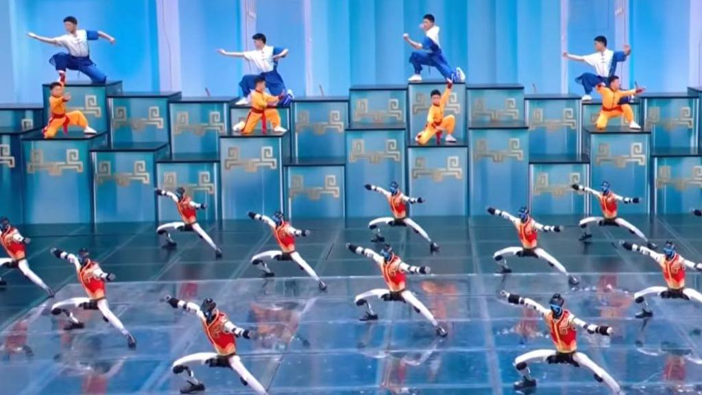 A screengrab shows robots in gold and red vests performing martial arts on stage during a Lunar New Year showcase, alongside young athletes carrying out a similar routine in the background.