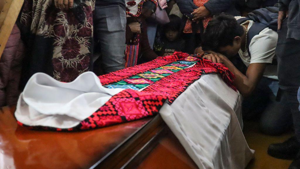 A man is hunched over the coffin of Father Marcelo Perez during his wake. On top of the coffin, the priest's garments are laid out. 