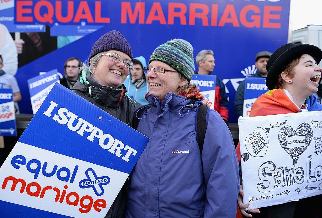 A group of women close to camera in cold weather jackets and woolly hats holding banners saying "I support equal marriage". They are laughing and cheering. There are men with similar banners in the background.