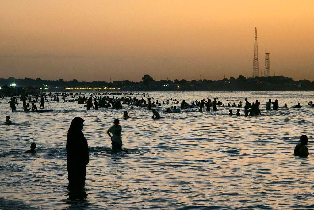 People swim in the sea at sunset. A woman wearing an abaya and hijab can be seen in the foreground standing in the water up to her ankles.