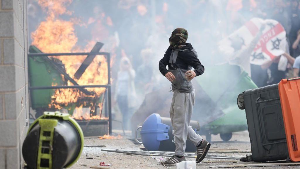 A masked figure wearing grey trousers and a dark top holds a stone while a skip burns in the background, creating smoke which covers a group of people during a riot.   Other objects have been thrown onto the ground.