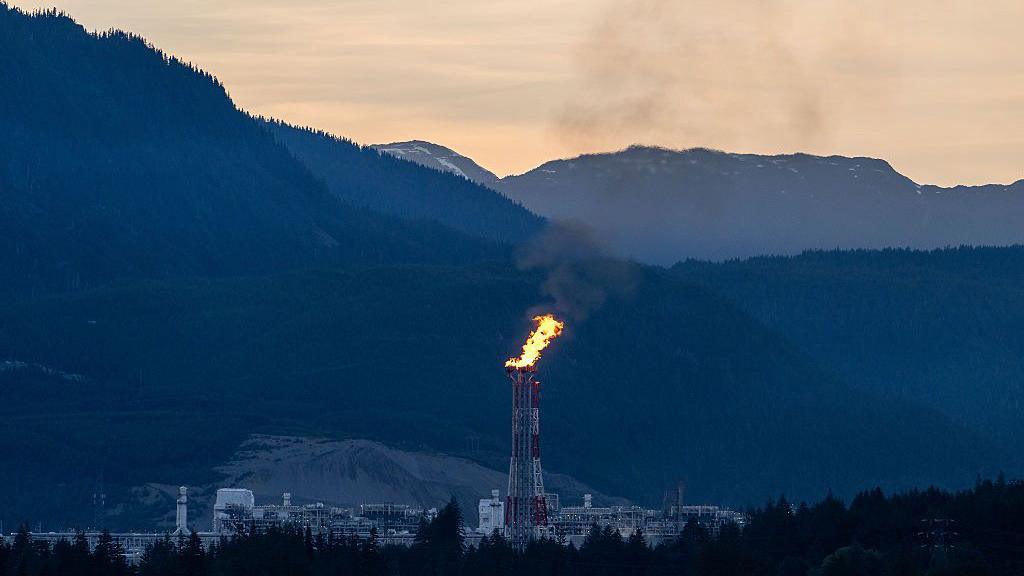 A white and red flare stack at sends fire up into the evening sky at the LNG Canada facility in Kitimat, British Columbia, Canada. In the background are mountains juxtaposed against a pale orange sky.