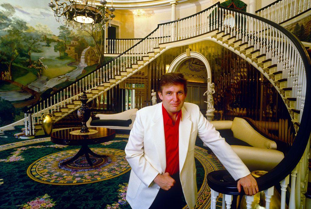 Donald Trump, dressed in a red shirt and white blazer, stands in a huge, grand hallway in his mansion.