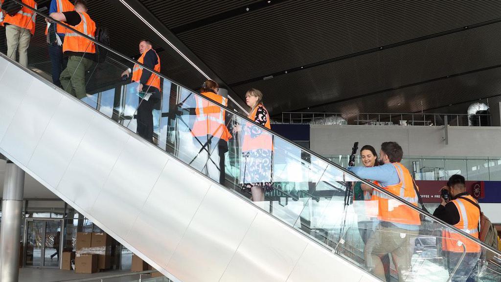 Several people wearing hi-vis vests stand on an escalator