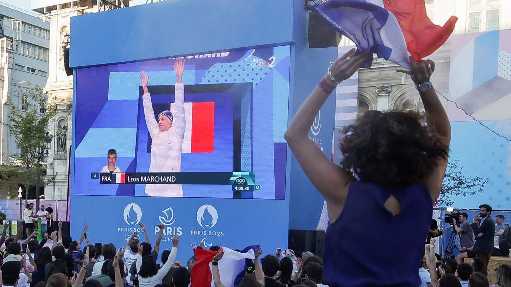 A woman in the foreground cheers as swimmer Léon Marchand appears on a big screen