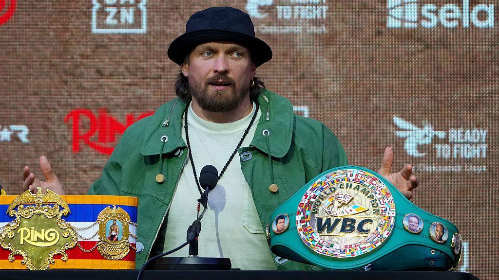 Oleksandr Usyk sits at a table behind the WBC and The Ring heavyweight titles