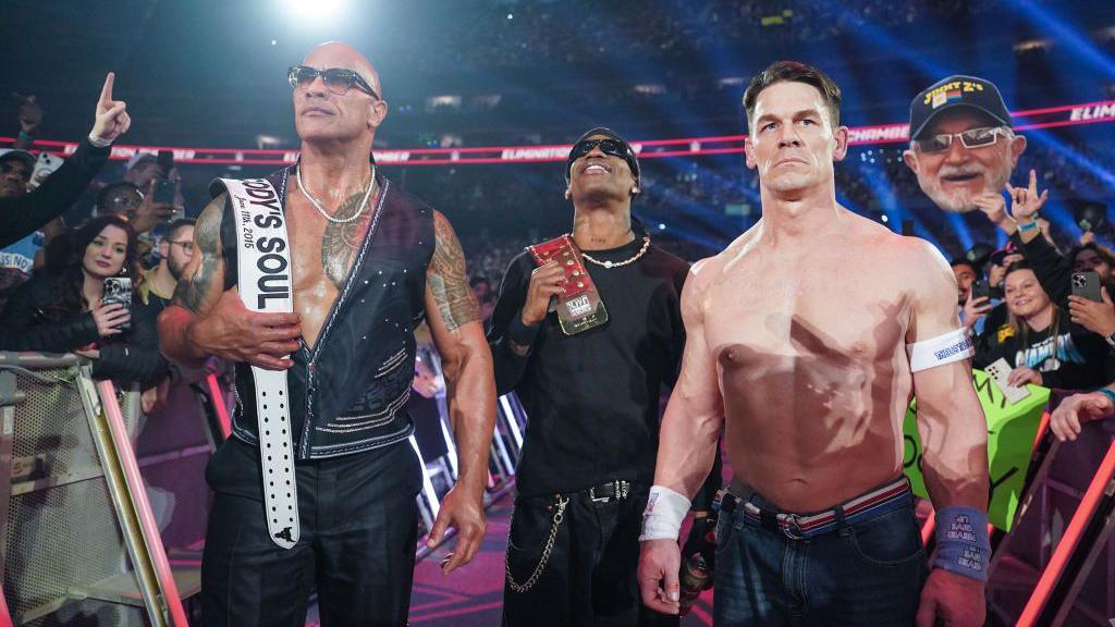 The Rock, a bald man wearing sunglasses and a waistcoat with nothing on with tattoos on his arms and torso is holding a belt which says Cody's Soul, just behind to the right is a man with a hat and sunglasses (Travis Scott) also holding a belt on his shoulder  and in front to the right is John Cena wearing jeans and no top. They are standing in an arena with crowd behind them. 