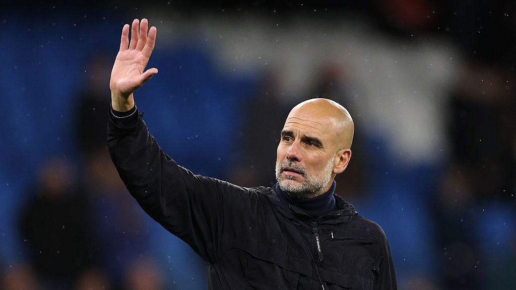 pep Guardiola wearing a black jacket and waving.