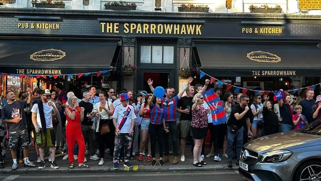 Crowd of people wearing Crystal Palace colours celebrating outside The Sparrowhawk pub in Crystal Palace