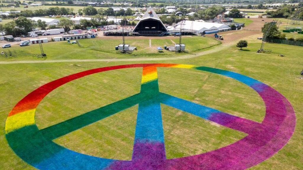 Huge rainbow peace sign on grass in front of the Pyramid Stage at Glastonbury Festival. There are several white tents in the background and lots of white vans.