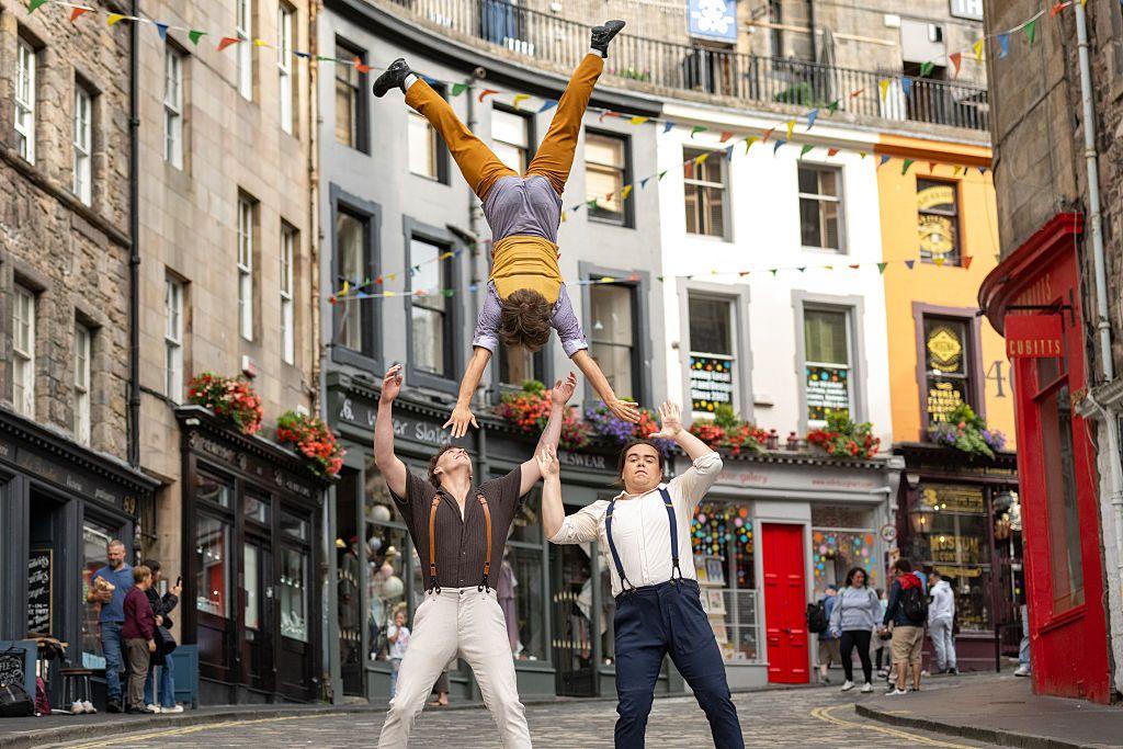 Three members of the cast of circus company Flip Fabrique perform a human pyramid in Victoria Street. They are wearing trousers with braces and are in front of the famous street with coloured buildings that curves behind them.