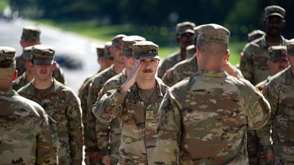 Several US military members in military uniform. In the centre, a man with Cladwell written on his name patch salutes a man in front of him. 