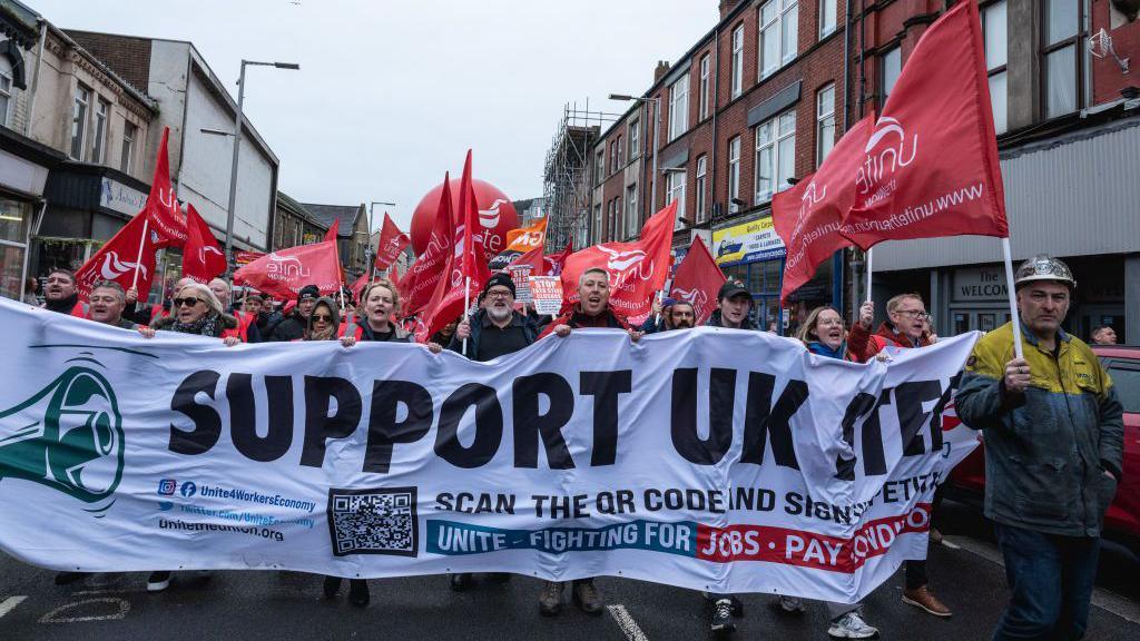 Protesters holding a white banner and red Unite flags. They are marching on a street. 