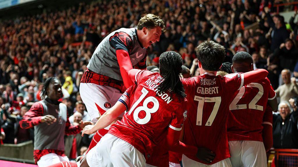 Charlton Athletic players celebrate in a huddle in front of fans sitting in the stands in the background