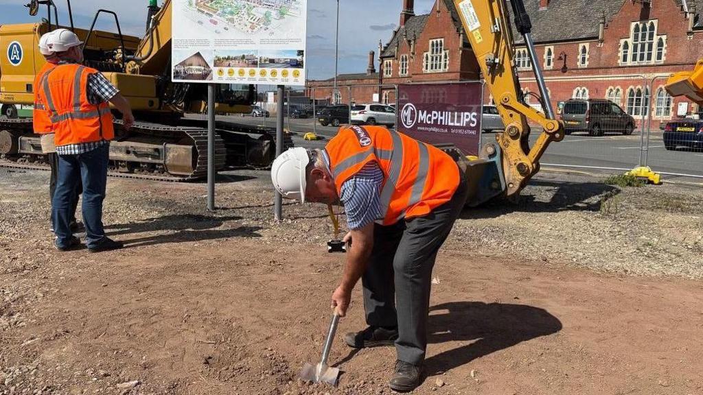 Two men wearing high-viz jackets, stood on a mound of soil - one of the men is bending over sticking a spade in the ground.