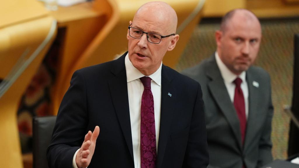 John Swinney in dark suit, white shirt and purple tie has his right hand slightly raised as he answers a question