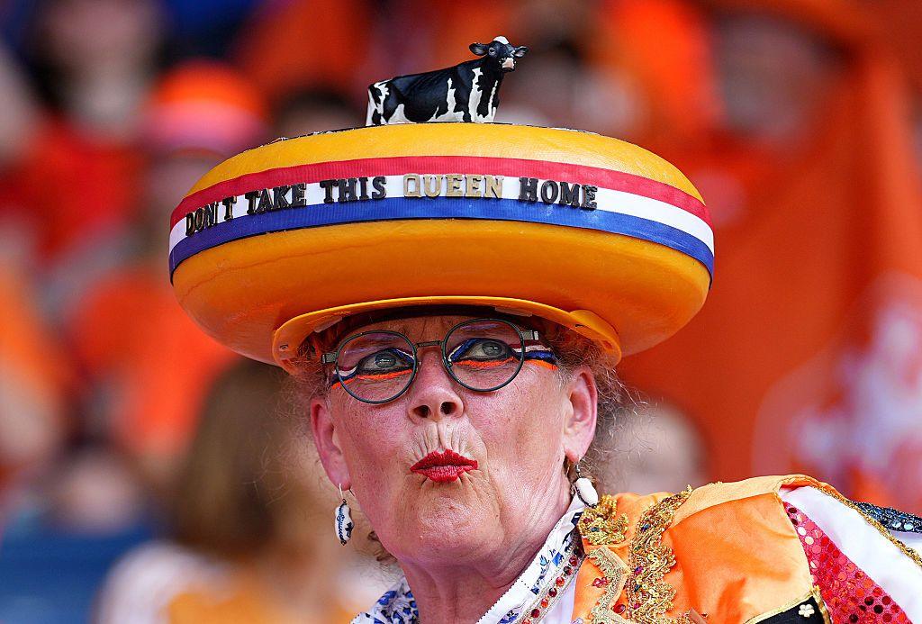 A middle-aged woman wears a hat resembling a Gouda cheese. The hat has a striped ribbon in the colours of the Netherlands flag displaying the words "Don't take this queen home". She is also wearing red lipstick and red, white, blue and orange eye make-up