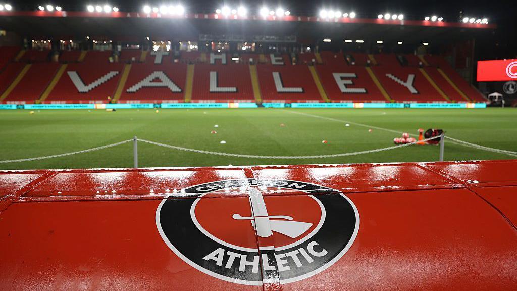 A view of Charlton Athletic's empty football stadium, The Valley, at night under bright floodlights. In the foreground, the top of a red dug-out seat or tunnel structure is prominently featured, displaying the circular Charlton Athletic crest with a hand holding a sword. Beyond the green pitch, red stadium seating spells out "The Valley" in large white lettering across the main stand.