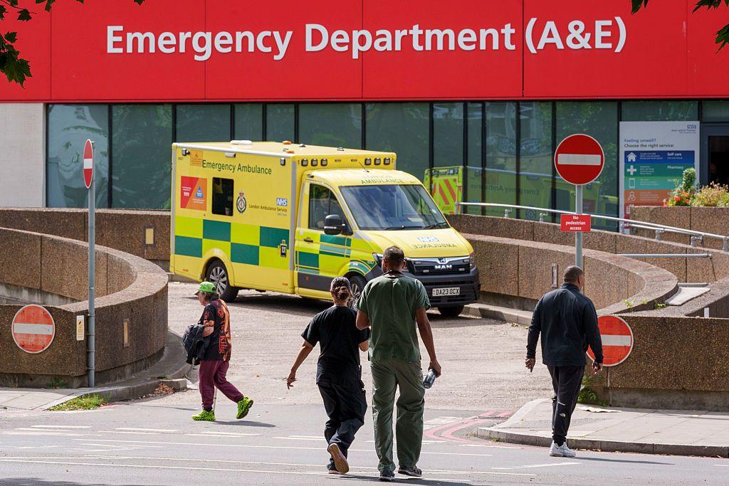 members of the public and two anonymous doctors walking outside an emergency department with an ambulance parked near the entrance.