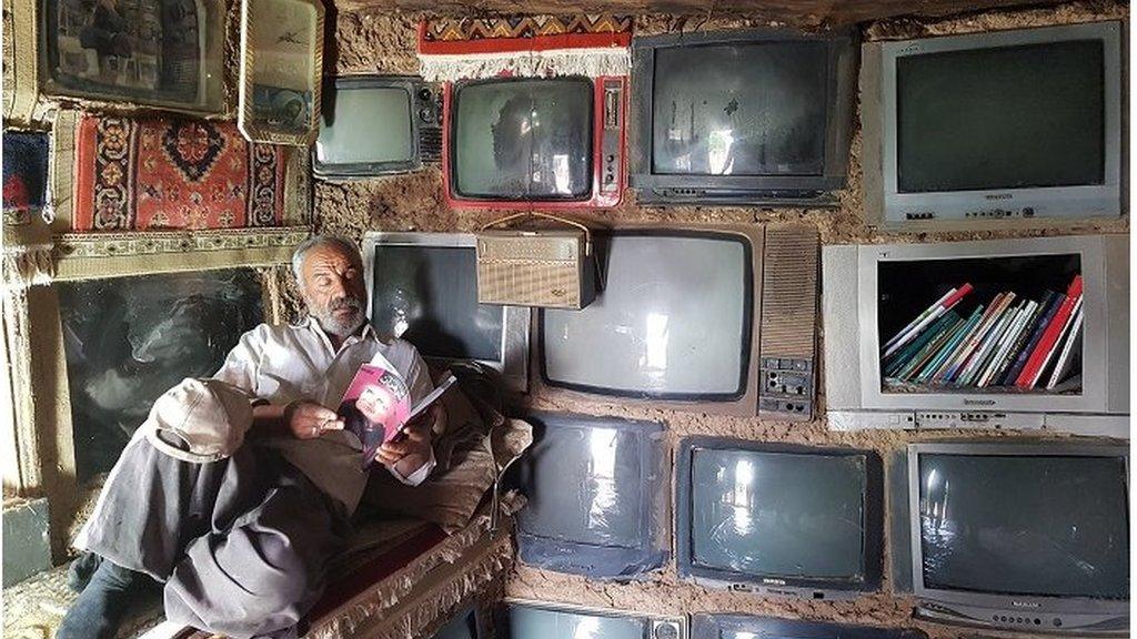 An image of a man sitting reading with a number of different televisions on the wall