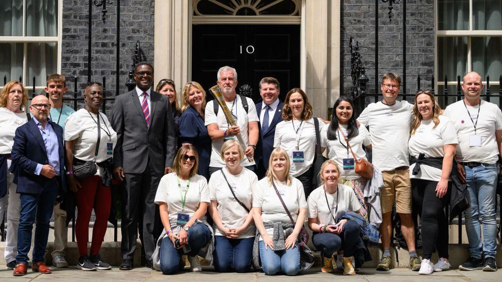 Former journalist Mike McCarthy holds the "Baton of Hope" as it is delivered to number 10, Downing Street following a tour of the country on July 06, 2023 in London, England. There are two lines of people sitting or kneeling for the photo