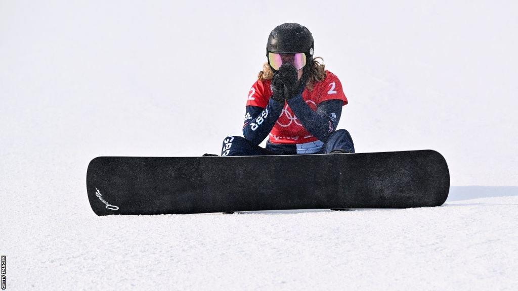 Charlotte Bankes sits on the snow, still strapped to her snowboard, with her hands over her mouth after being knocked out of the Olympics in Beijing in 2022