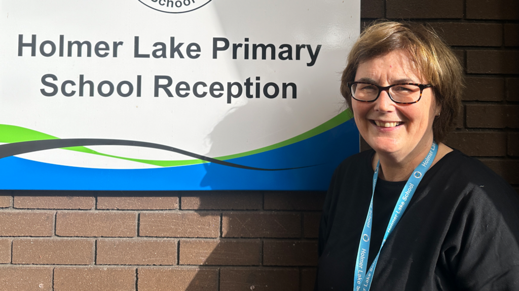 A woman with brown hair, glasses and a dark top, wearing a blue lanyard and standing in front of a white sign which reads Holmer Lake Primary School reception