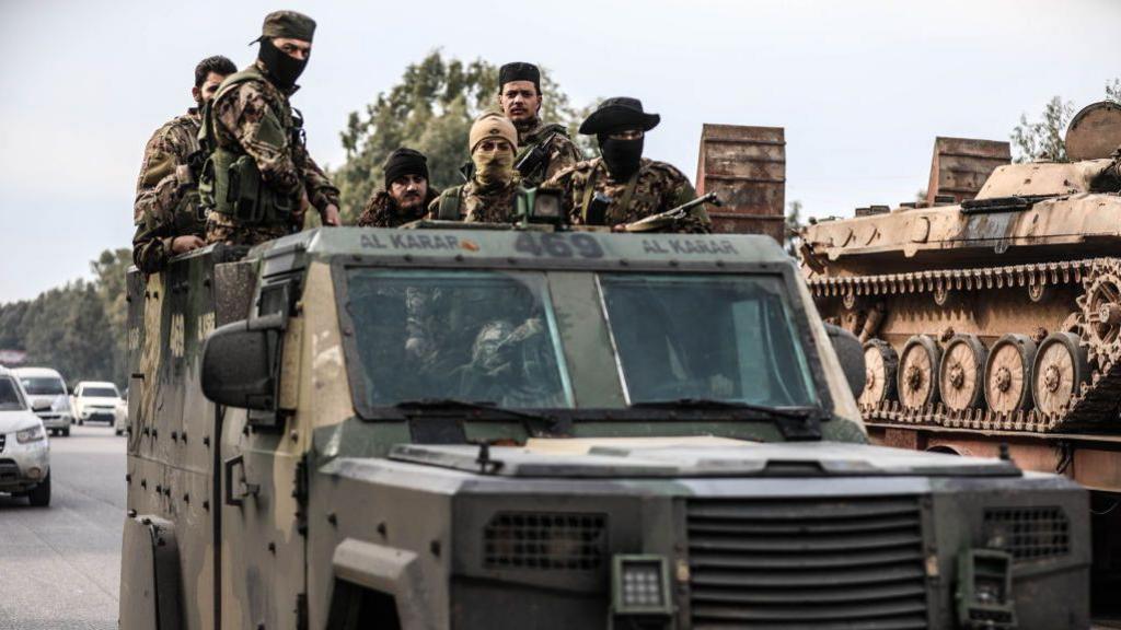 Armoured vehicle with six soldiers standing in the back of it. Four are wearing hats and three are wearing face coverings. The vehicle is on a road and in the background there is a tank.