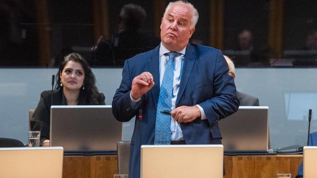 Andrew RT Davies, wearing a blue suit, tie and shirt, asking a question in the Senedd chamber watched by Conservative colleague Natasha Asghar