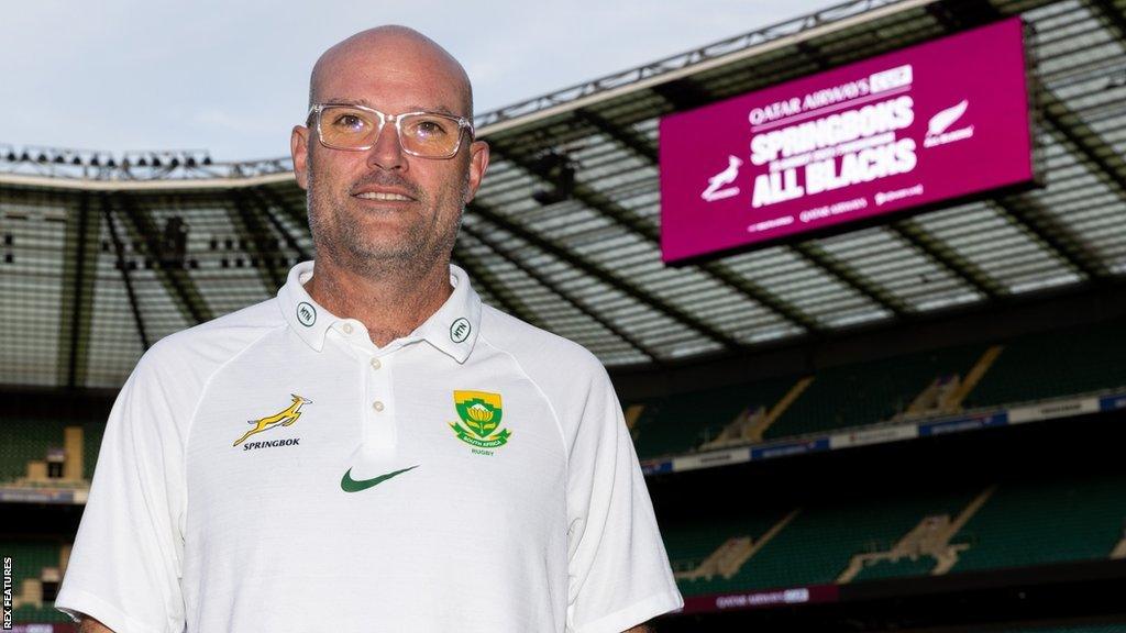 Jacques Nienaber pitchside at Twickenham standing below a scoreboard advertising the match between South Africa and New Zealand