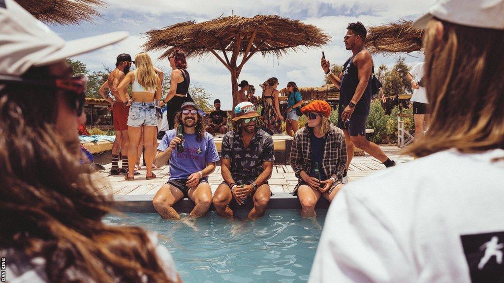 A group of runners sit on the edge of a swimming pool drinking beers