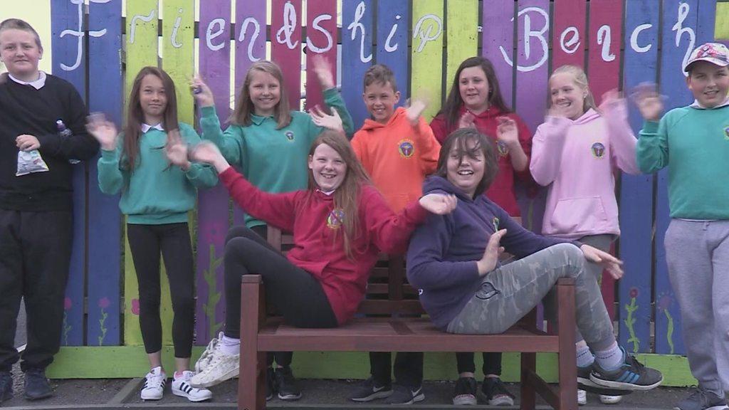 A group of children sitting on and around a bench with the words friendship bench painted on a fence behind them